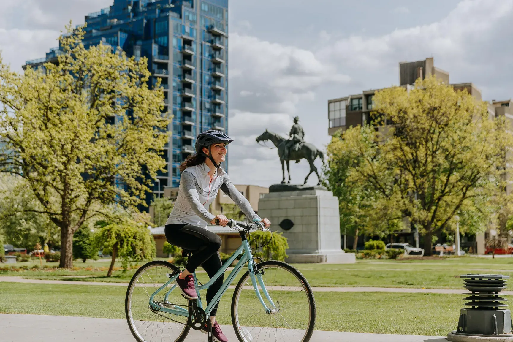 Woman on a bicycle rides past an equestrian statue and modern buildings in a green city park.