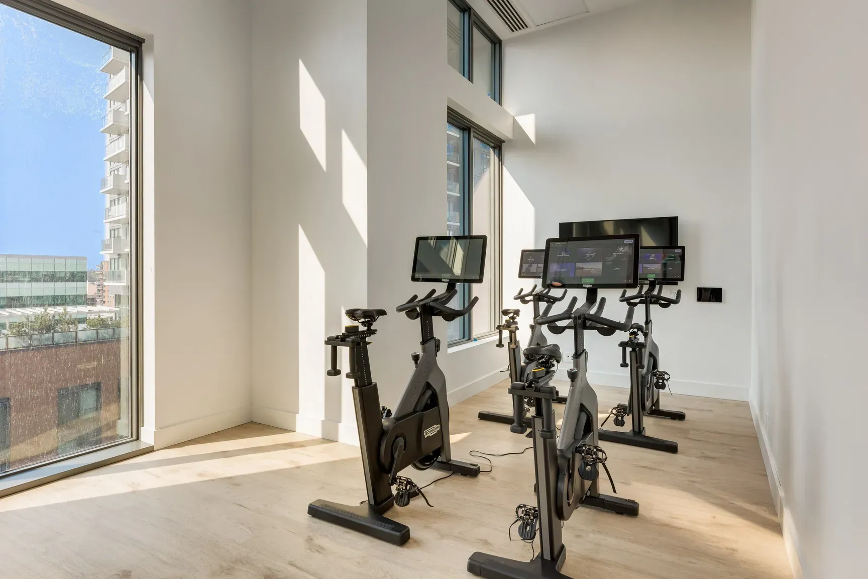 Several exercise bikes with screens in a bright, modern gym room with large windows.