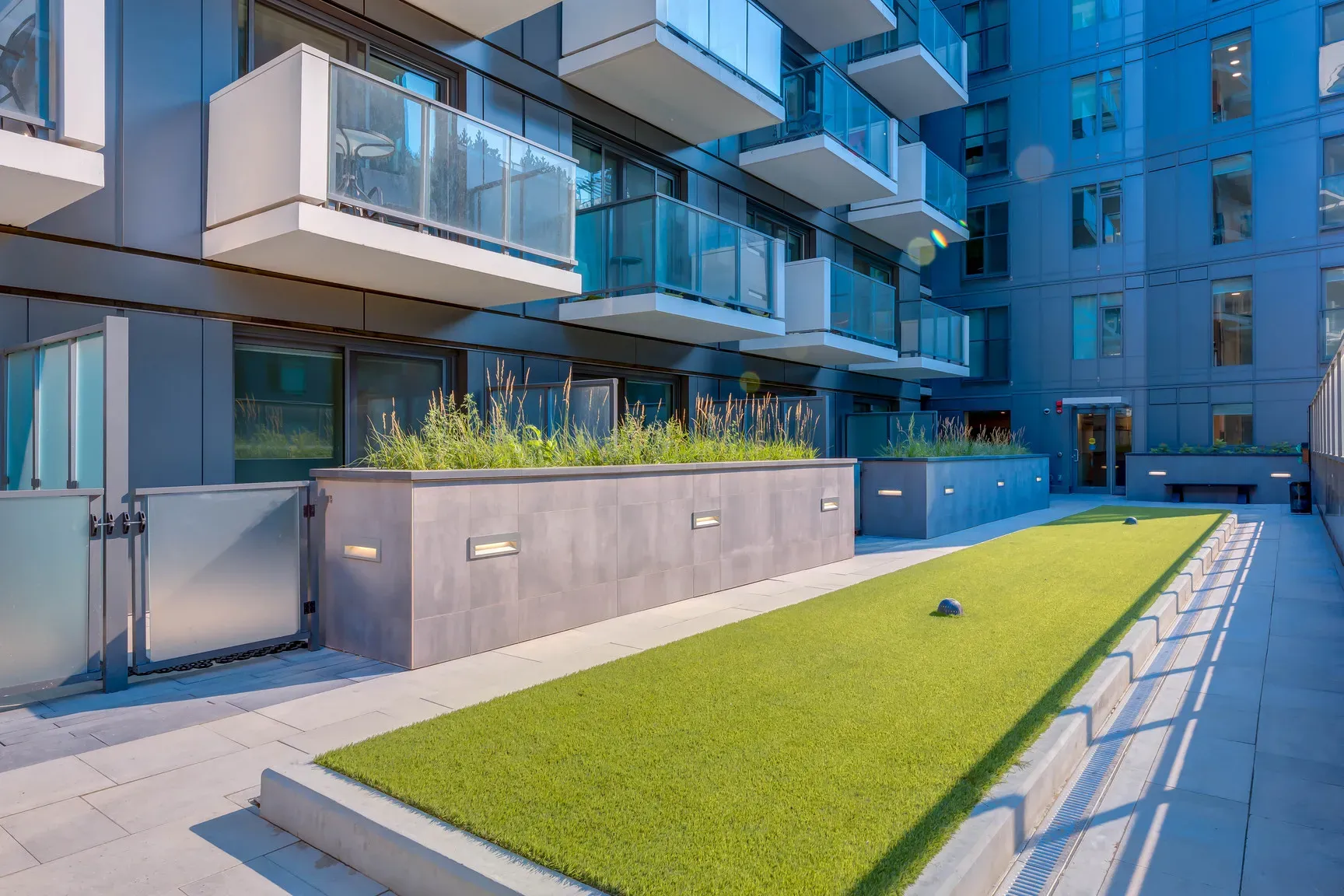 Modern building with glass balconies overlooking an artificial putting green and concrete planters.